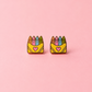 A pair of small enamel earrings shaped like yellow crayon boxes with colorful crayons on top and a pink heart on the front, shown on a solid pink background.