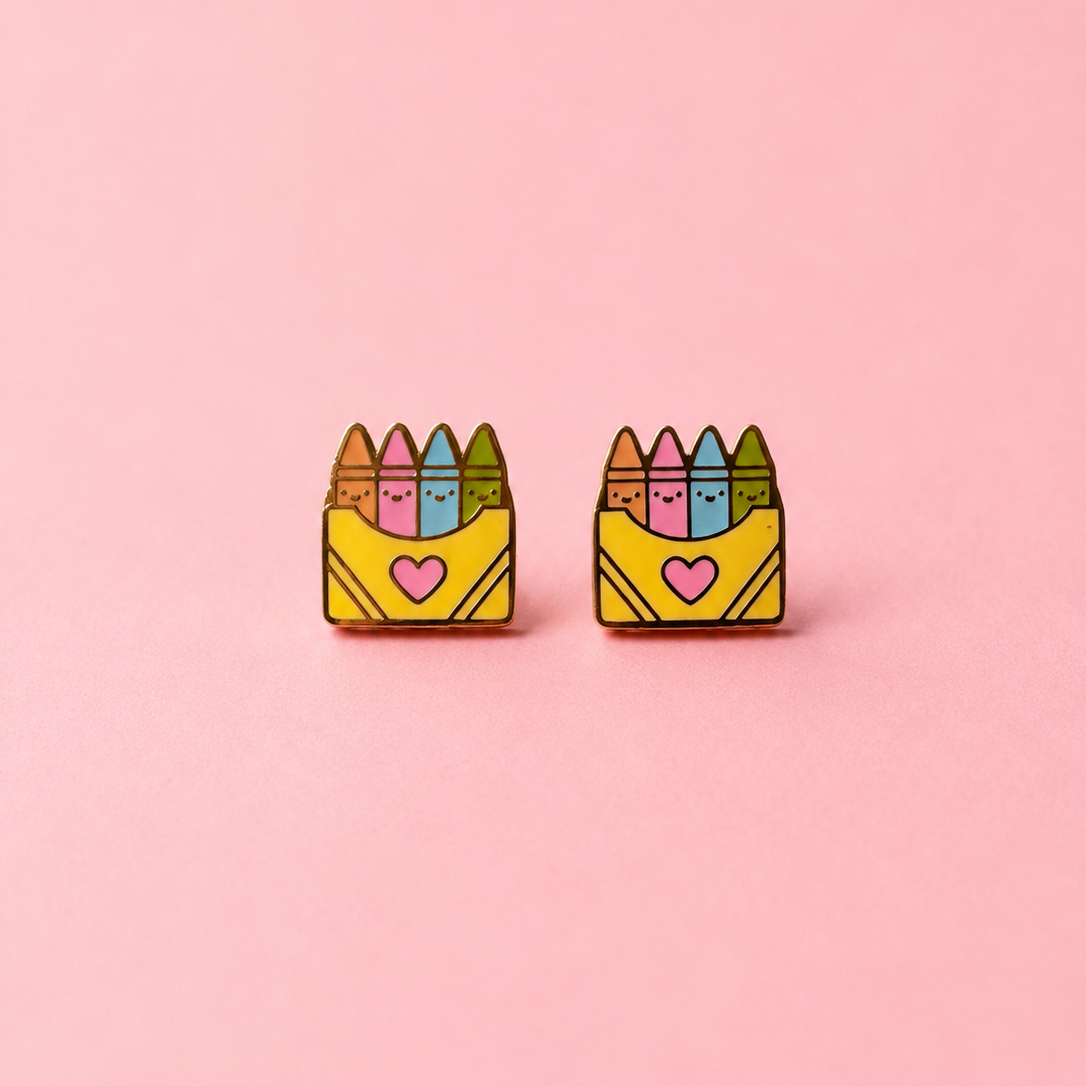 A pair of small enamel earrings shaped like yellow crayon boxes with colorful crayons on top and a pink heart on the front, shown on a solid pink background.