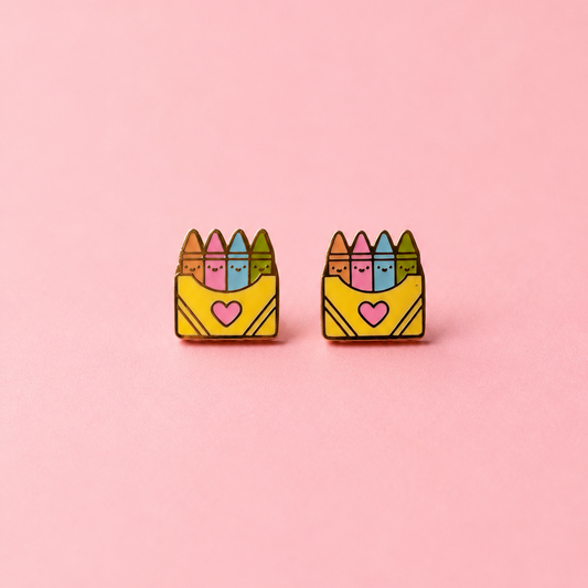 A pair of small enamel earrings shaped like yellow crayon boxes with colorful crayons on top and a pink heart on the front, shown on a solid pink background.