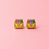 A pair of small enamel earrings shaped like yellow crayon boxes with colorful crayons on top and a pink heart on the front, shown on a solid pink background.