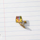 A gold-tone earring shaped like a yellow envelope holding colorful crayons, placed on a white lined notebook page.