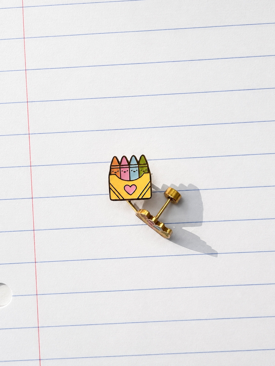 A gold-tone earring shaped like a yellow envelope holding colorful crayons, placed on a white lined notebook page.