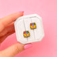 A hand with pink manicured nails holds a small white jewelry display card showing two matching enamel earrings. Each earring depicts a yellow crayon box with colorful crayons and a small pink heart, set against a solid pink background.