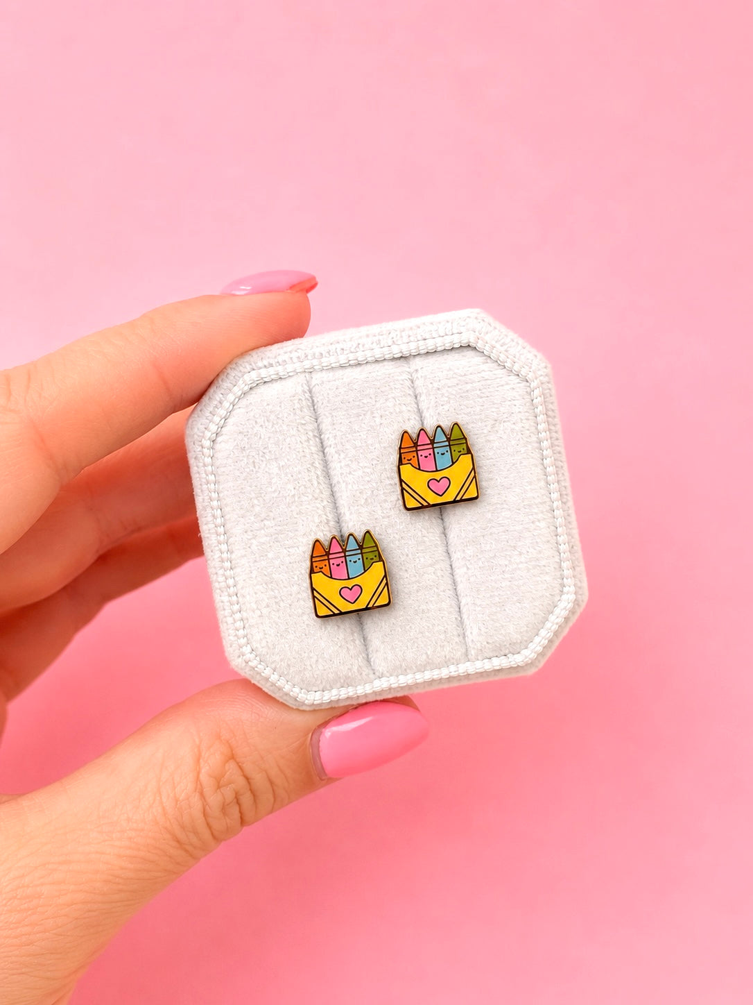 A hand with pink manicured nails holds a small white jewelry display card showing two matching enamel earrings. Each earring depicts a yellow crayon box with colorful crayons and a small pink heart, set against a solid pink background.