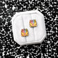 Two small enamel earrings shaped like yellow crayon boxes with colorful crayons and pink hearts, displayed on a white fabric octagonal stand against a black-and-white speckled background.