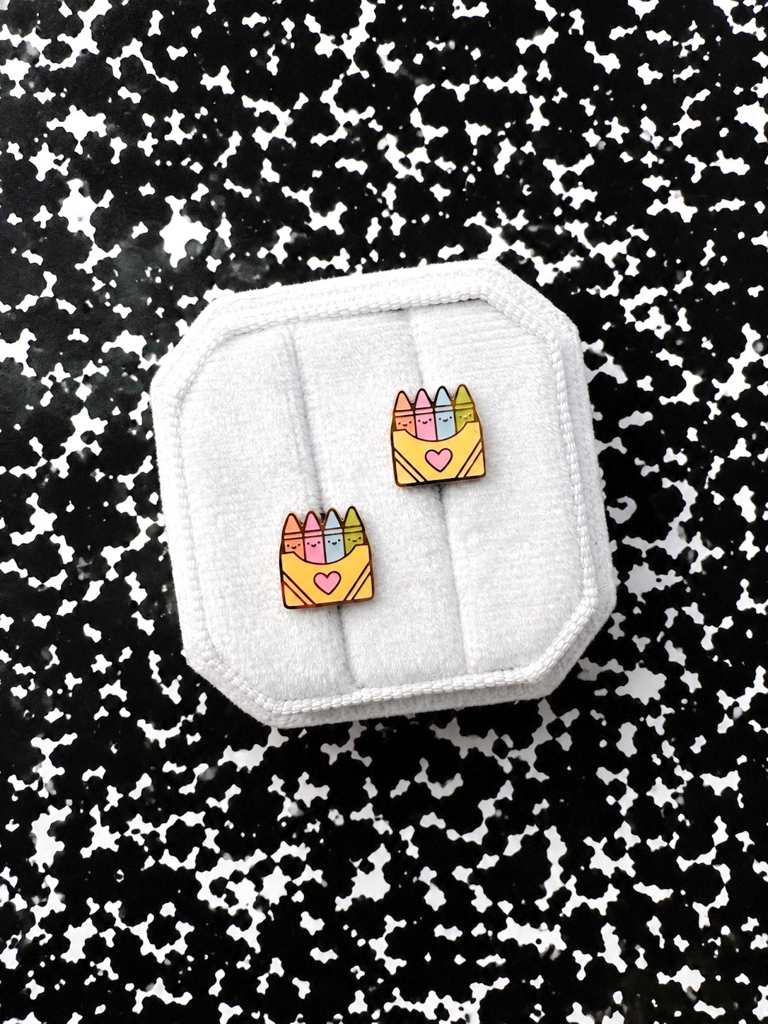 Two small enamel earrings shaped like yellow crayon boxes with colorful crayons and pink hearts, displayed on a white fabric octagonal stand against a black-and-white speckled background.