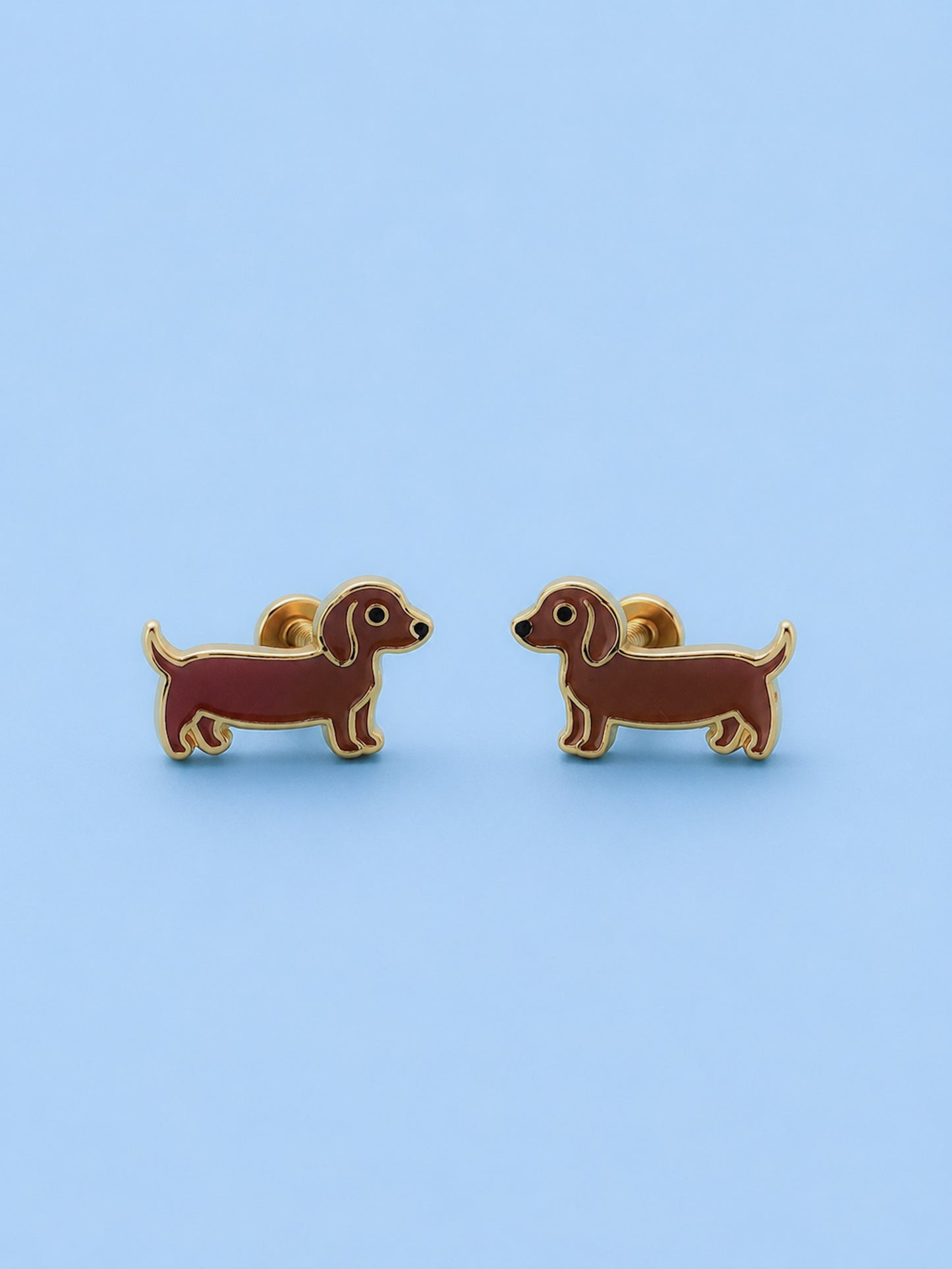 A pair of dachshund-shaped stud earrings in brown enamel with gold-tone metal outlines, displayed on a light blue background.