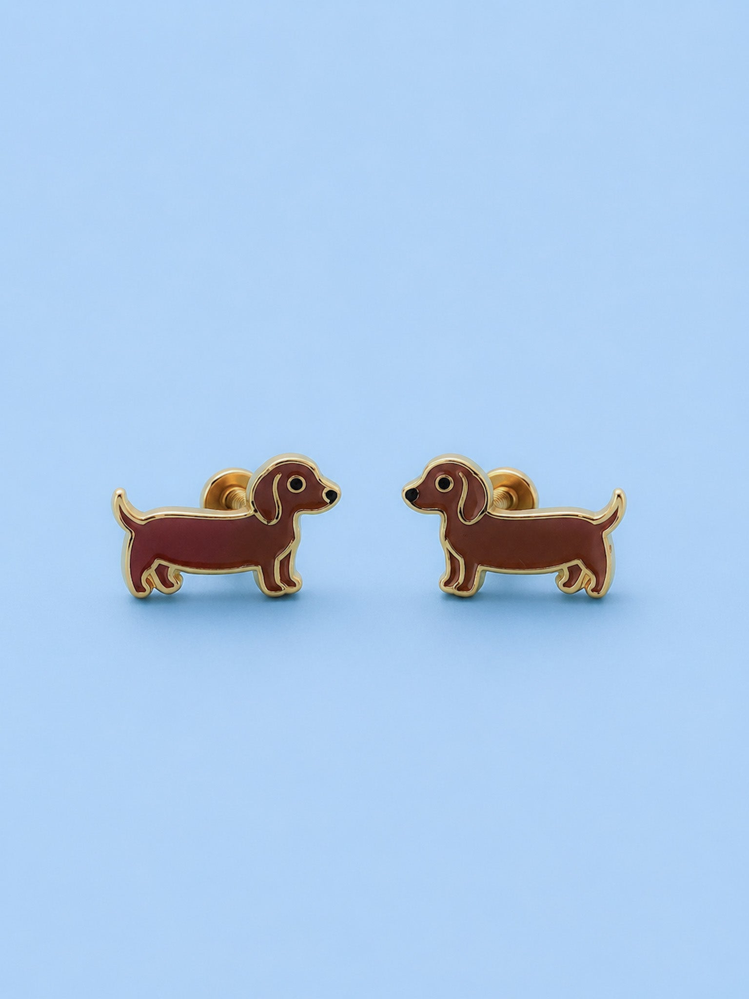 A pair of dachshund-shaped stud earrings in brown enamel with gold-tone metal outlines, displayed on a light blue background.