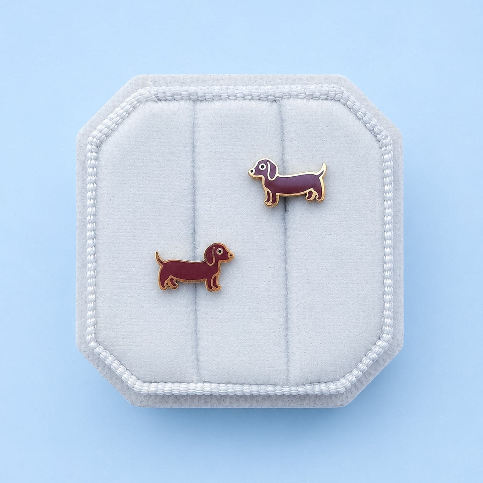 A pair of small dachshund-shaped stud earrings in dark brown and gold-tone metal, displayed on a white octagonal jewelry box against a light blue background.