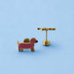 A pair of gold-tone earrings shaped like a pink dachshund dog, shown on a solid light blue background. One earringk is facing forward to show the dog profile, and the other is turned sideways to show the post and flat-back clasp.