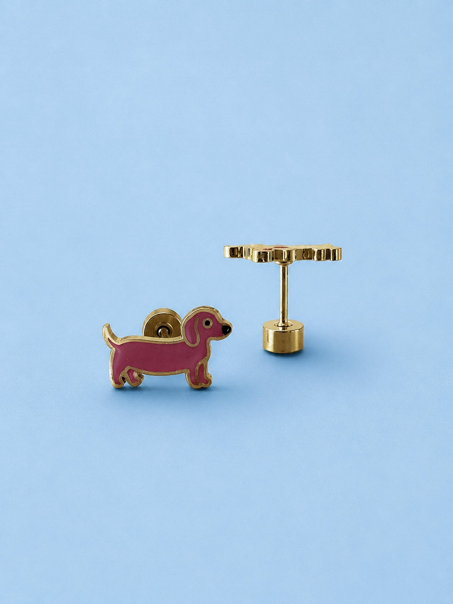 A pair of gold-tone earrings shaped like a pink dachshund dog, shown on a solid light blue background. One earringk is facing forward to show the dog profile, and the other is turned sideways to show the post and flat-back clasp.