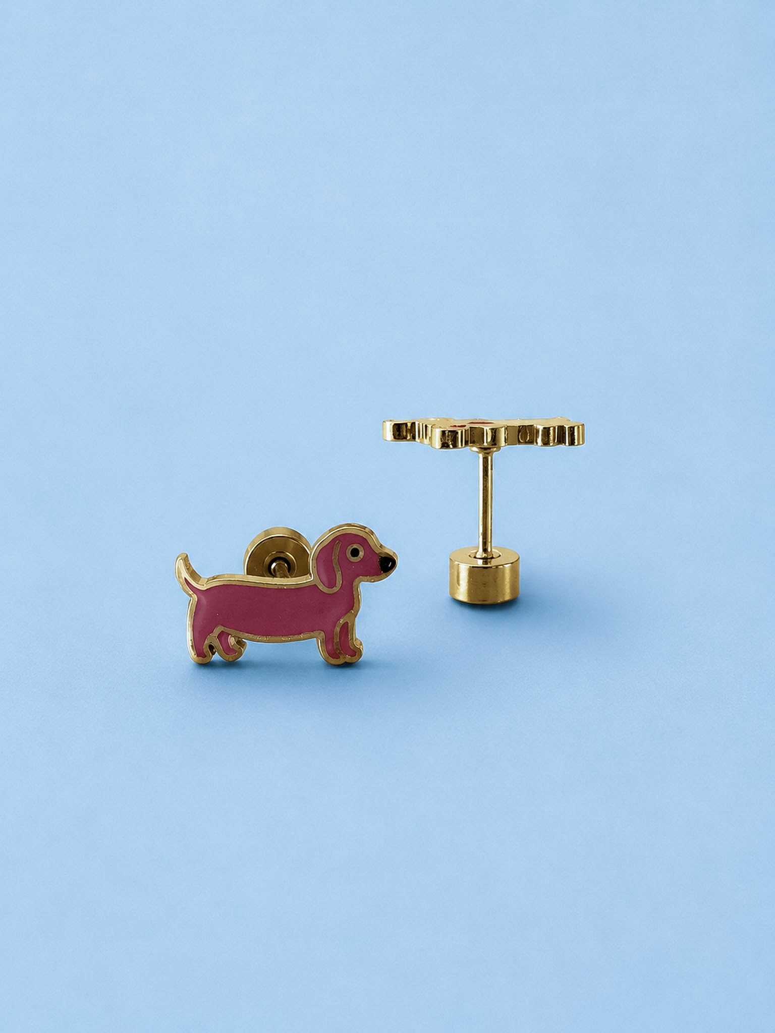 A pair of gold-tone earrings shaped like a pink dachshund dog, shown on a solid light blue background. One earringk is facing forward to show the dog profile, and the other is turned sideways to show the post and flat-back clasp.