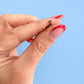 A close-up of a hand with red French-tipped nails holding a small gold earring shaped like a dachshund against a solid light blue background.
