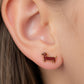 A close-up of a person's ear wearing a small dachshund-shaped stud earring in dark brown enamel with gold-tone outlines.