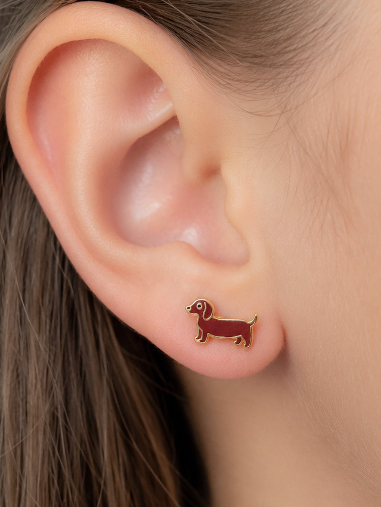 A close-up of a person's ear wearing a small dachshund-shaped stud earring in dark brown enamel with gold-tone outlines.