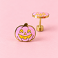 A pair of gold-tone earrings shaped like jack-o'-lantern pumpkins, featuring a pink glitter enamel face with yellow details, set against a solid pink background.
