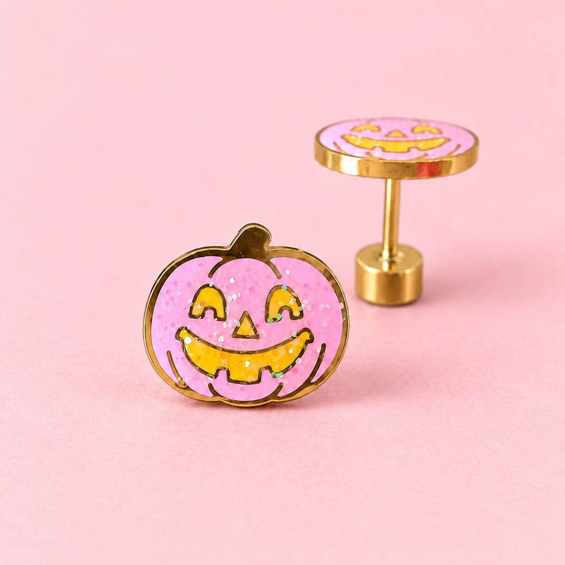 A pair of gold-tone earrings shaped like jack-o'-lantern pumpkins, featuring a pink glitter enamel face with yellow details, set against a solid pink background.