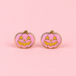 A pair of pumpkin-shaped stud earrings with a pink glittery enamel face, yellow jack-o'-lantern facial features, and gold-tone metal borders, displayed on a solid pink background.