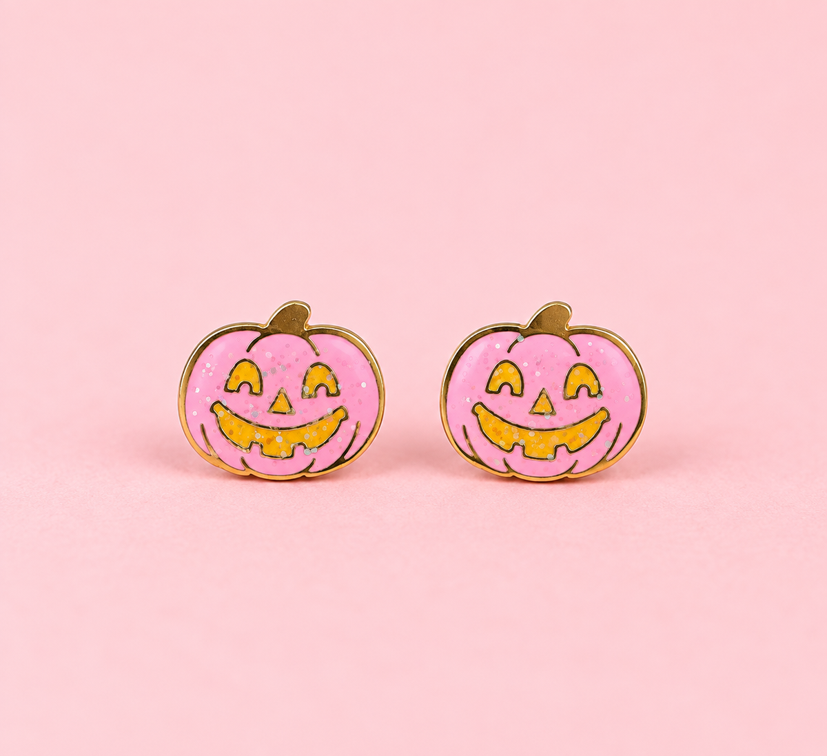 A pair of pumpkin-shaped stud earrings with a pink glittery enamel face, yellow jack-o'-lantern facial features, and gold-tone metal borders, displayed on a solid pink background.