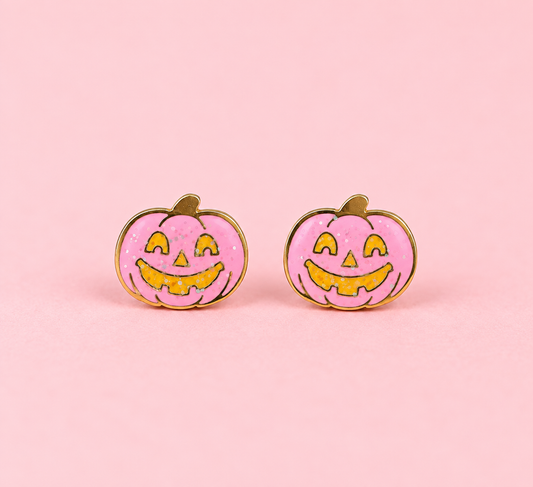 A pair of pumpkin-shaped stud earrings with a pink glittery enamel face, yellow jack-o'-lantern facial features, and gold-tone metal borders, displayed on a solid pink background.