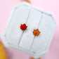 Two maple leaf earrings on a white card with a pink background
