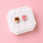 Two milk carton shaped stud earrings. One chocolate, one strawberry in a white jewelry box on a light pink background