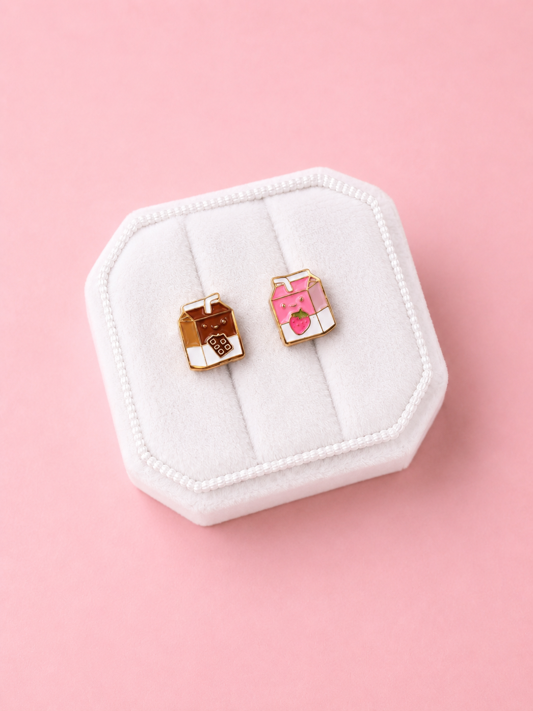 Two milk carton shaped stud earrings. One chocolate, one strawberry in a white jewelry box on a light pink background