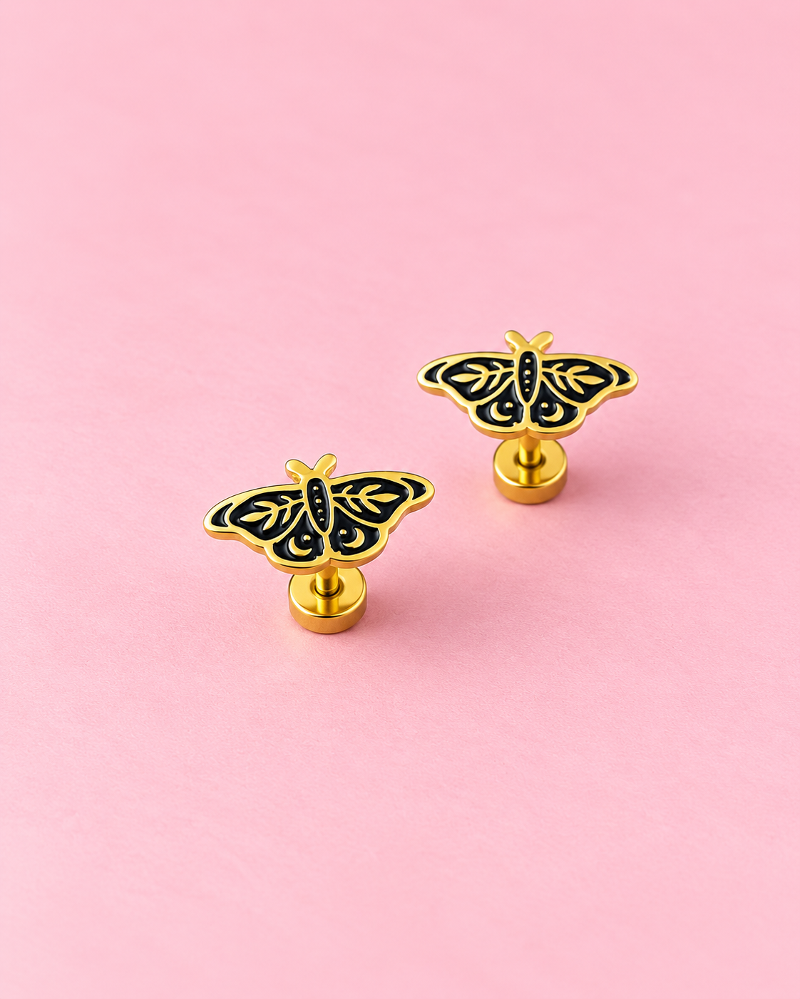 A pair of gold butterfly-shaped stud earrings with black enamel detailing, displayed on a solid pink background.