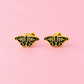 A pair of gold butterfly stud earrings with black enamel detailing, displayed on a solid pink background.
