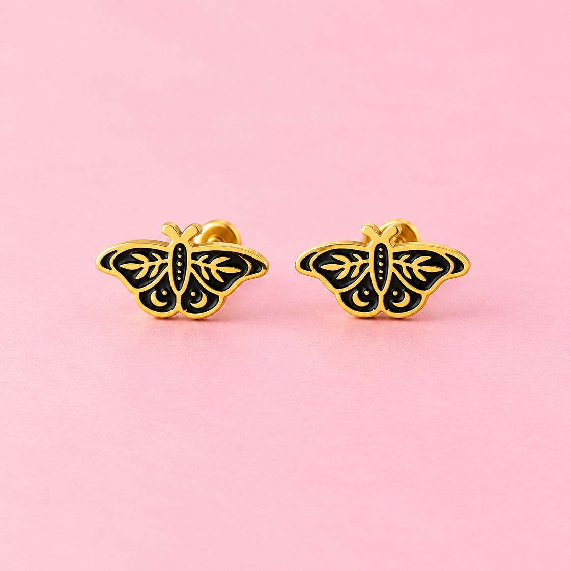 A pair of gold butterfly stud earrings with black enamel detailing, displayed on a solid pink background.