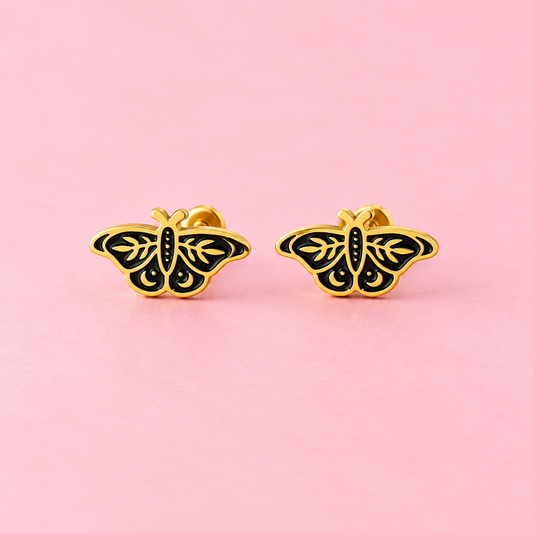A pair of gold butterfly stud earrings with black enamel detailing, displayed on a solid pink background.