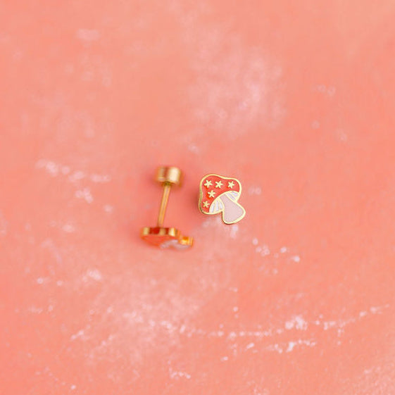 Two small gold mushroom earrings on a pink textured background