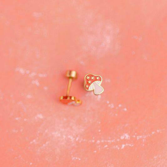 Two small gold mushroom earrings on a pink textured background