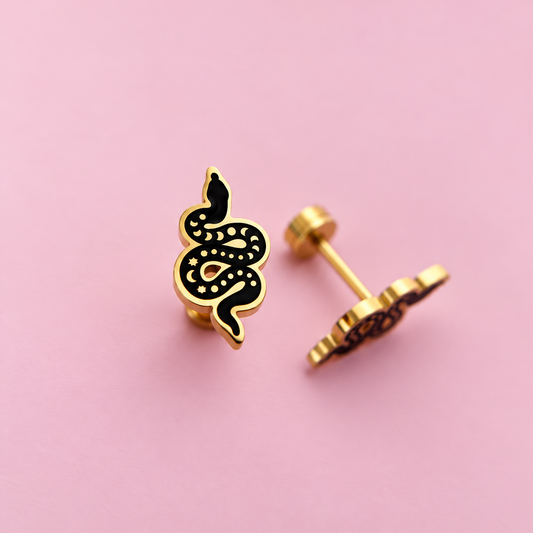 Gold snake stud earrings with black enamel detailing, shown from both front and side angles on a solid pink background.