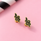 A pair of gold and black snake stud earrings displayed on a light pink background with a blurred black and white striped prop in the top left corner.