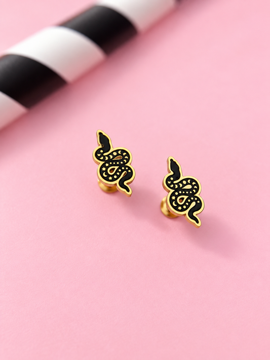 A pair of gold and black snake stud earrings displayed on a light pink background with a blurred black and white striped prop in the top left corner.