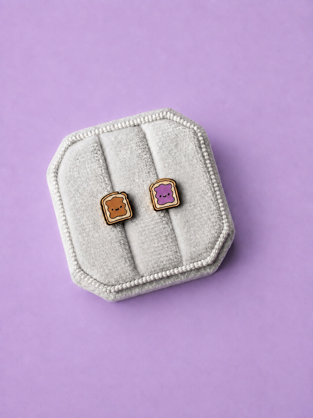 Peanut butter and jelly shaped enamel stud earrings in a white jewelry box on a light purple background
