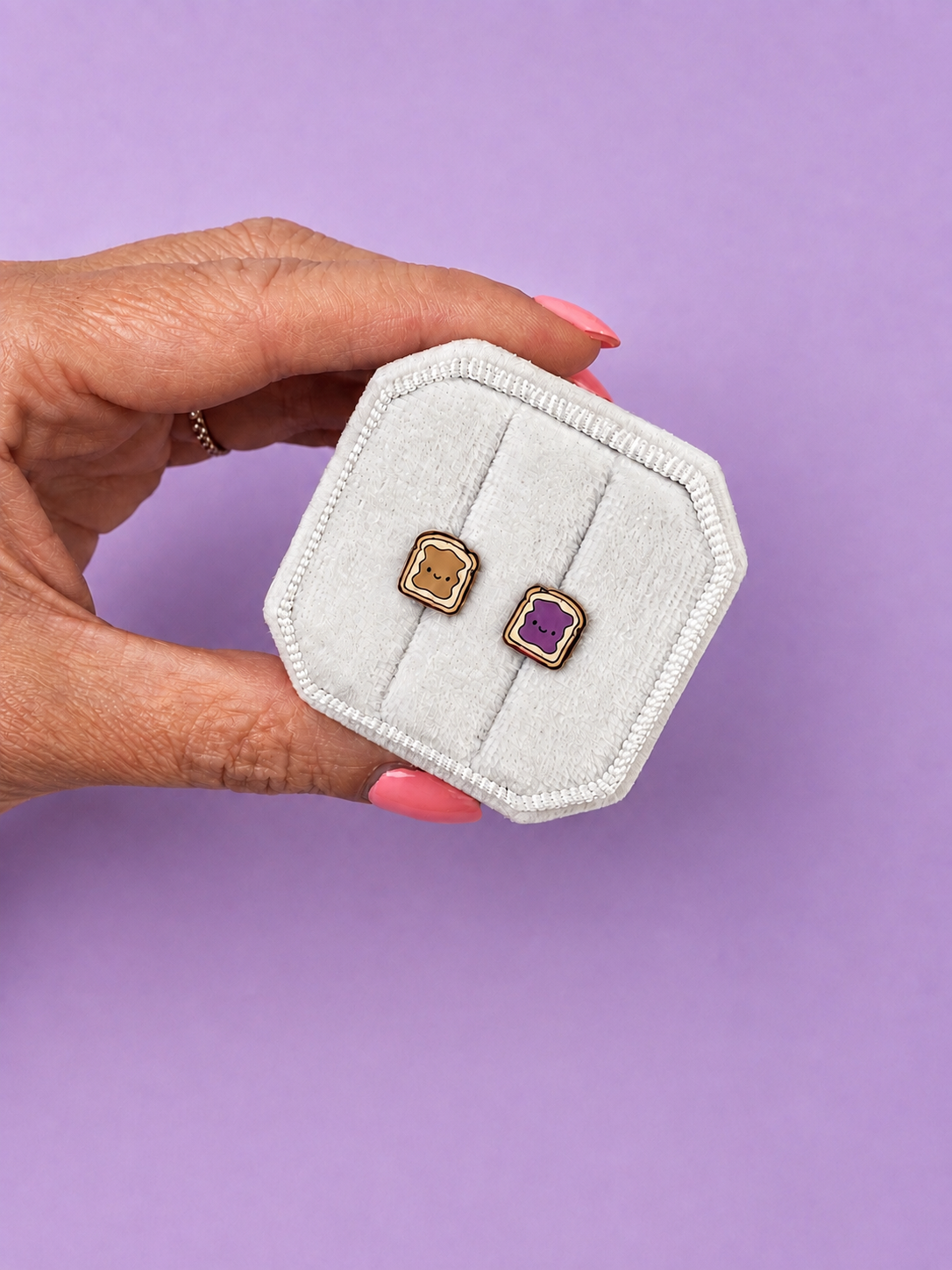Hand holding a white jewelry box with Peanut butter and jelly shaped enamel stud earrings on a light purple background