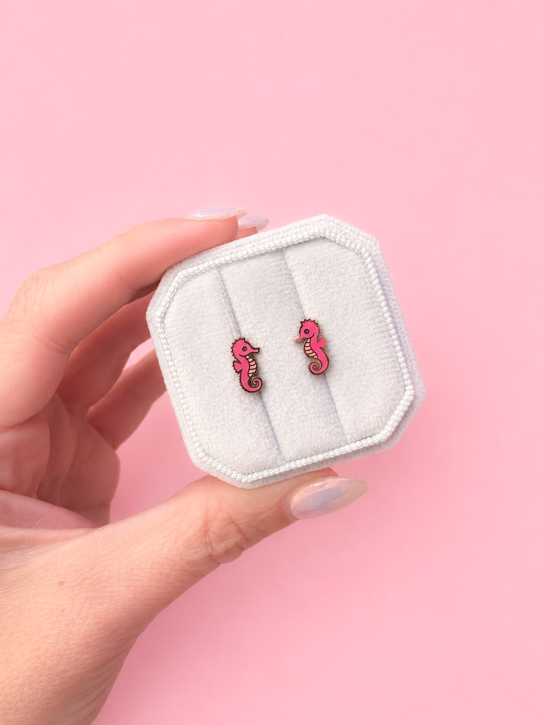 Hand holding a small white box with pink seahorse earrings against a pink background