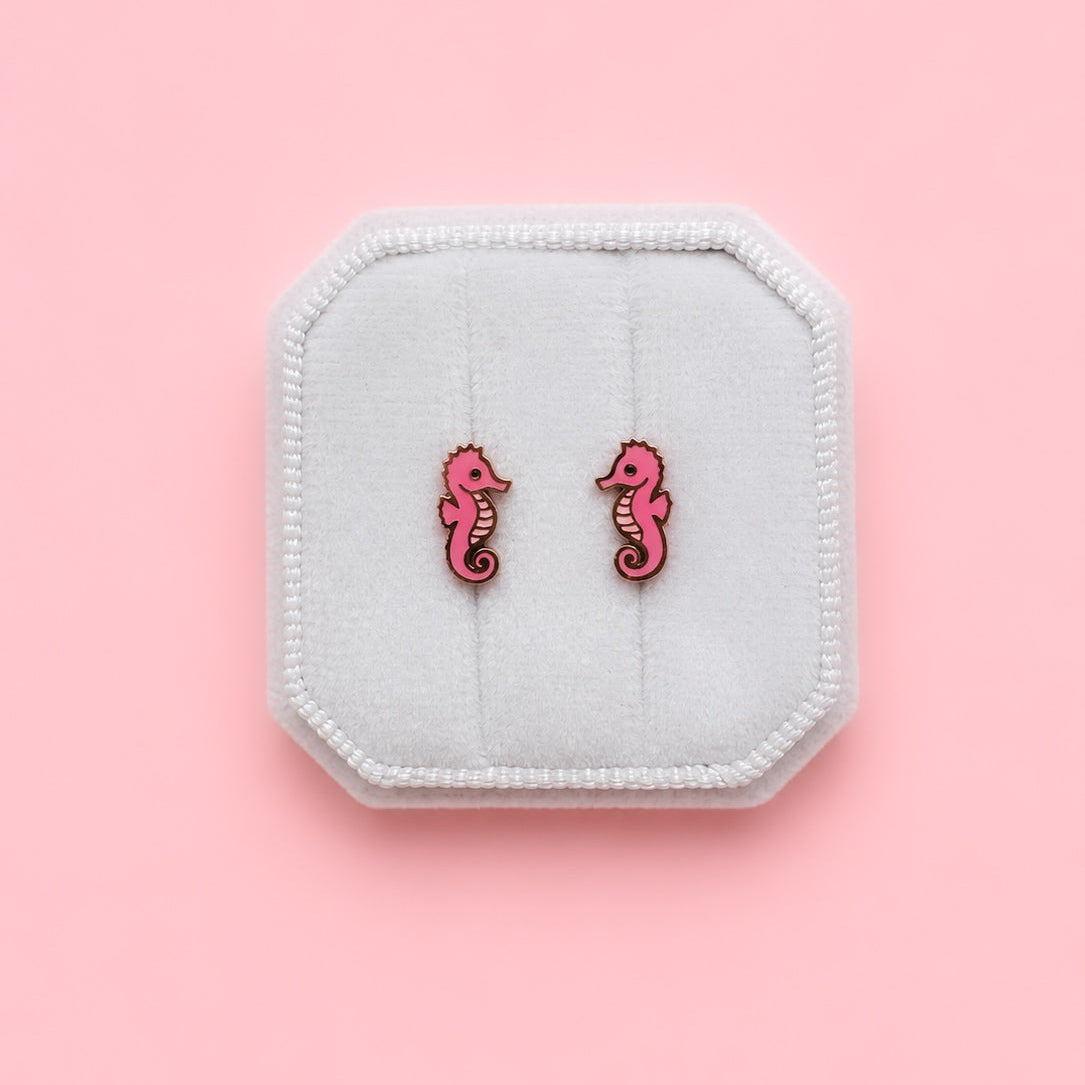 Pink seahorse earrings on a white jewelry box against a pink background