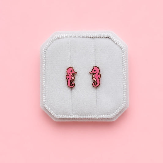 Pink seahorse earrings on a white jewelry box against a pink background