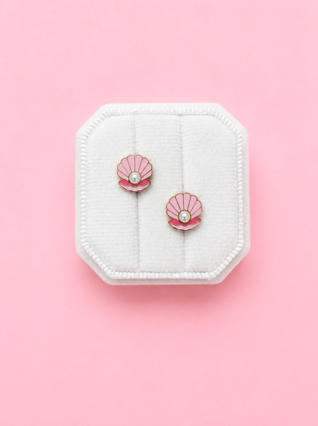 Pair of shell-shaped earrings with pearls on a white jewelry box against a pink background