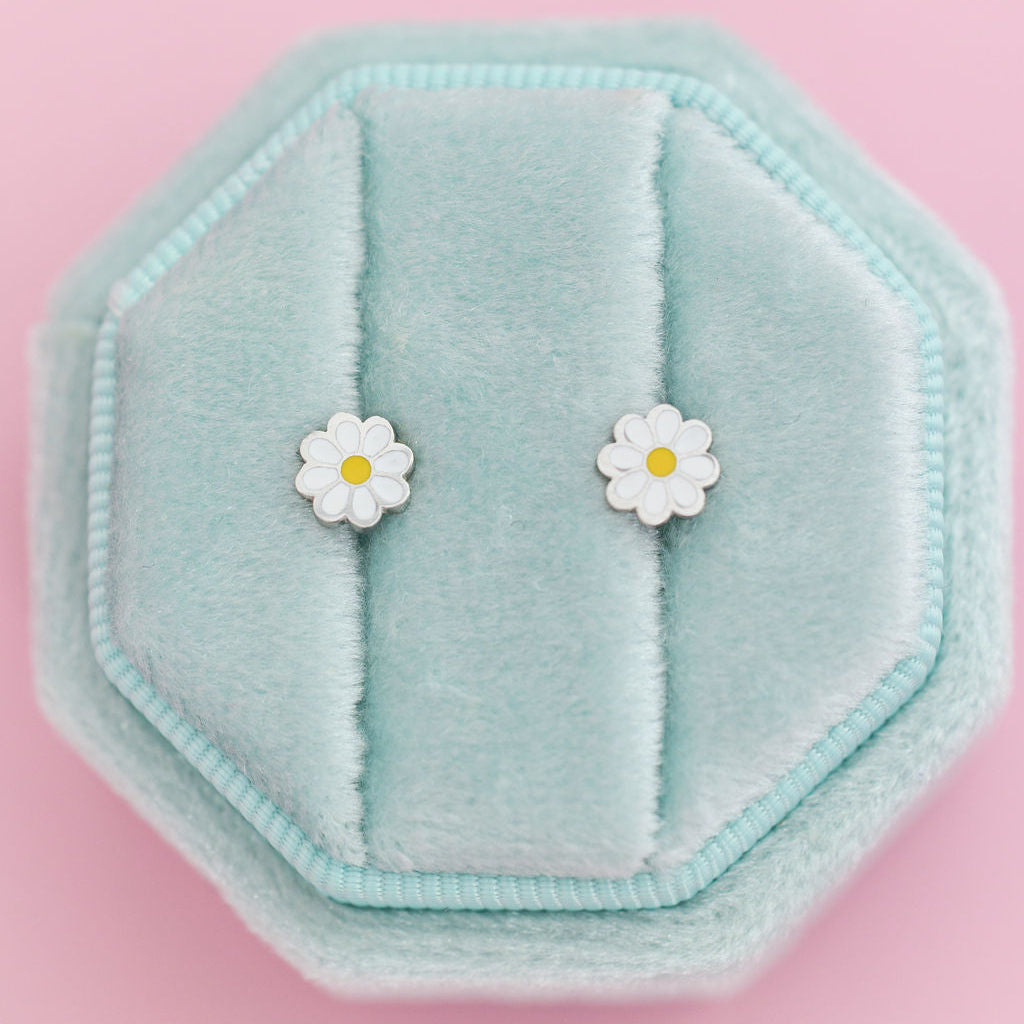 A pair of small daisy stud earrings with white petals and yellow centers, displayed on a mint blue octagonal jewelry box against a solid pink background.