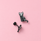 A small black and white skeleton cat stud earring shown next to its black post and backing on a solid pink background.