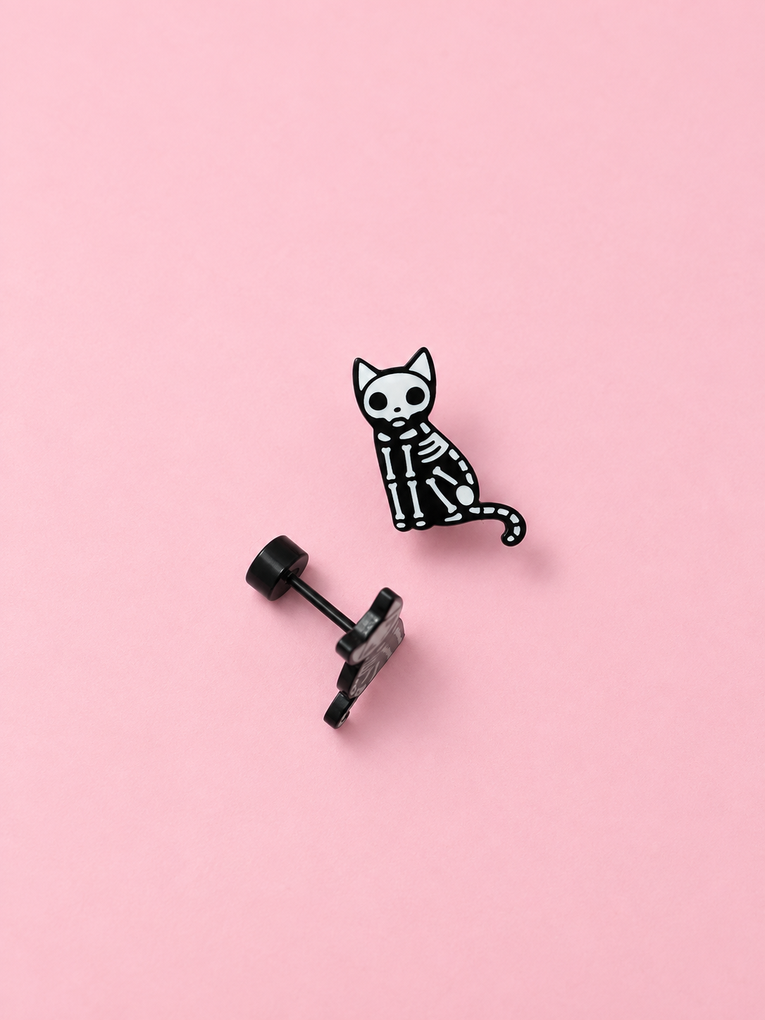 A small black and white skeleton cat stud earring shown next to its black post and backing on a solid pink background.