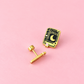 A gold-tone stud earring with an enamel book pin featuring a crescent moon and the word 'SPELLS' on a pink background.