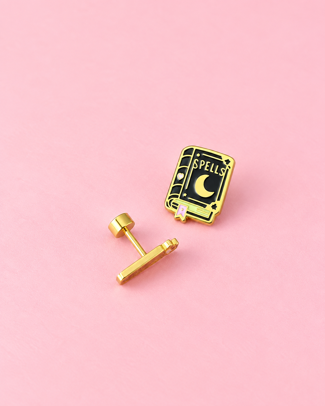 A gold-tone stud earring with an enamel book pin featuring a crescent moon and the word 'SPELLS' on a pink background.