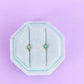 Pair of earrings in a hexagonal blue holder on a light purple background