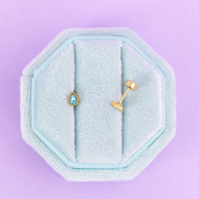 Two gold earrings in a light blue hexagonal earring holder on a purple background