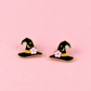 A pair of witch hat stud earrings with black enamel, gold metal trim, a small pink flower, and a gold crescent moon, displayed on a solid pink background.